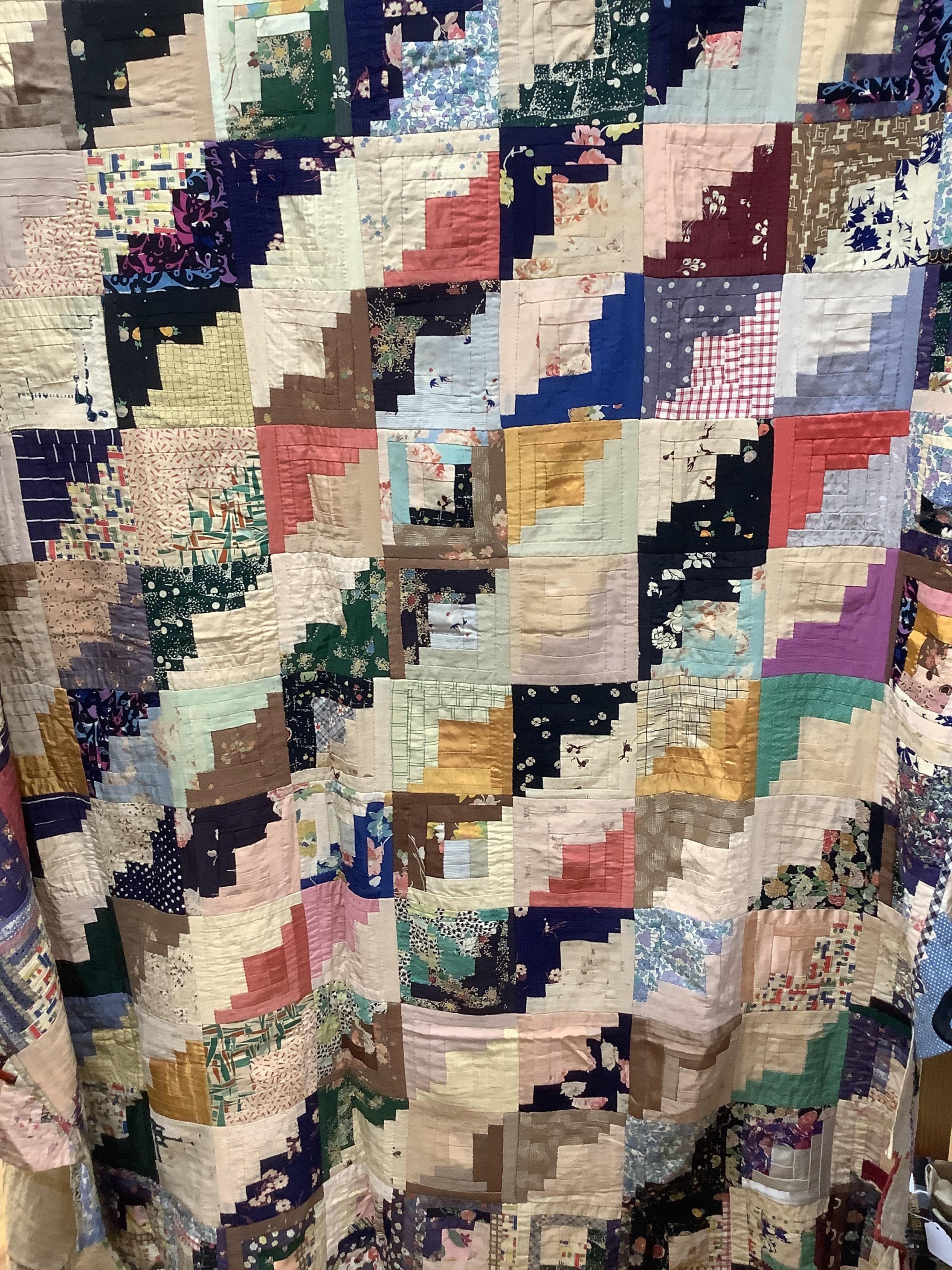 An early 20th century Log Cabin patchwork bedcover, worked in plain and printed silk, rayons and cottons from 1900 to 1940’s. Edged with a triangular patchwork border on three sides. Cover unlined. 158cm wide x 192cm lon